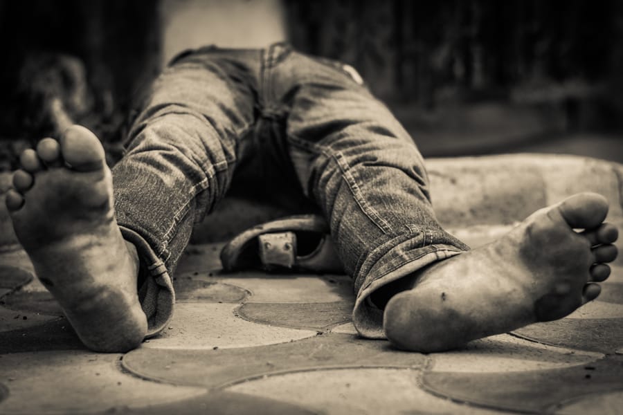 Alcoholic Anonymous. A man face-down in the gutter, drunk, mid-afternoon — bare feet, worn jeans, the warm stone holding him. Not a verdict, just a man at his lowest, set down here as plainly and as gently as the frame allows.