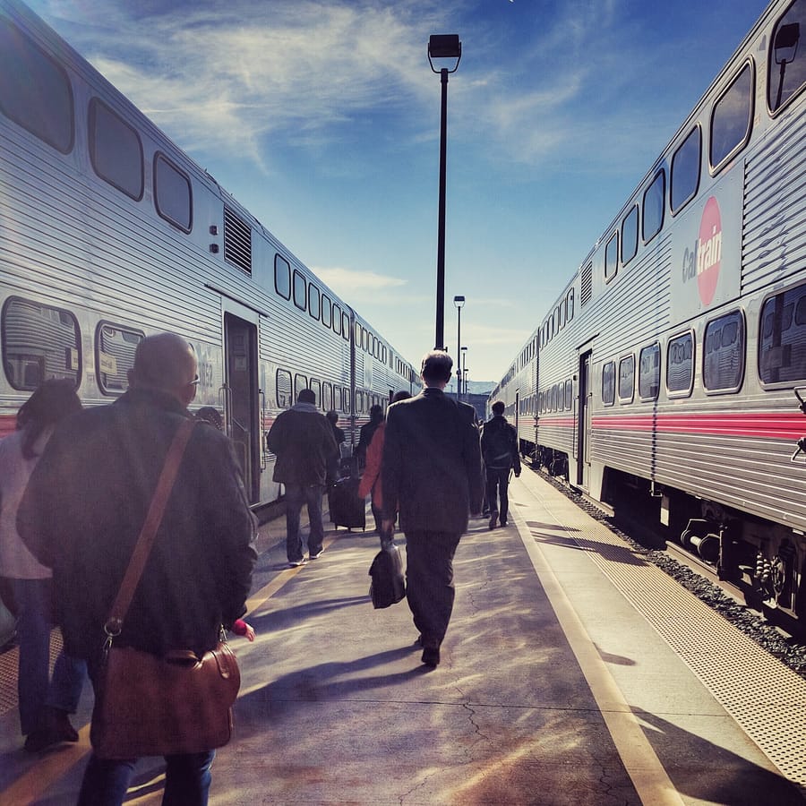 Between Trains. Commuters cross a Caltrain platform between two waiting trains, low winter sun raking the concrete. California.