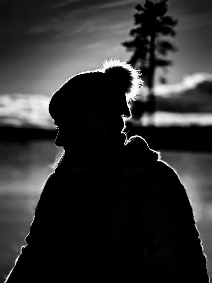 Silhouette at Dusk. A figure in a bobble hat against a rim-lit winter sky, a lone pine and still water behind. Drawn entirely in edges and backlight. Black and white.